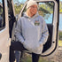 A woman wearing a light gray hoodie with a 'Somewhere Life & Free' graphic and a cream beanie, sitting in the open door of a white vehicle outdoors near water and trees.