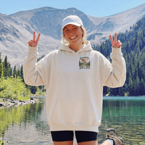 A woman wearing a cream-colored hoodie with a 'Summit Life & Free' graphic on the left chest, posing outdoors in front of a mountain and lake backdrop.