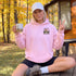 A woman sitting outdoors on a wooden deck in front of a cabin, wearing a light pink hoodie with a 'Sourpuss' graphic on the left chest, a white baseball cap, and black shorts, while holding a mug.