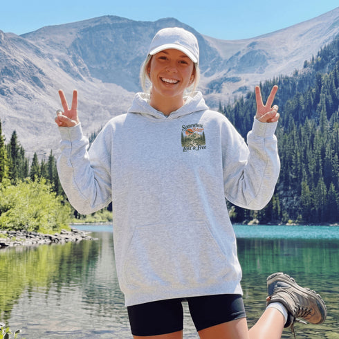 A woman stands outdoors in front of a mountain and lake, wearing a light heather gray hoodie with a small chest graphic that reads 'Sunrise Lake & Free,' a white cap, and black shorts. She is making peace signs with both hands.
