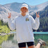 A woman stands outdoors in front of a mountain and lake, wearing a light heather gray hoodie with a small chest graphic that reads 'Sunrise Lake & Free,' a white cap, and black shorts. She is making peace signs with both hands.