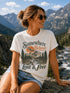 Woman wearing a t-shirt with a nature-themed design and text, sitting in a mountainous landscape.