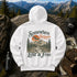A white pullover hoodie laid flat on a large rock, showing a scenic mountain graphic with the words 'Somewhere Between Lost & Free.' The hoodie is styled outdoors with a backpack, black cap, water bottle, map, compass, and hiking boots against a mountain landscape background.