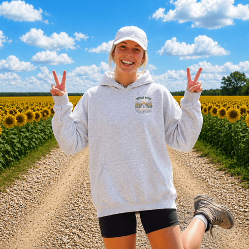 A woman stands on a dirt road between rows of sunflowers at sunset, wearing an oversized white hoodie with a small 'Wander where the sunflowers grow' chest logo and a white cap.