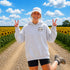 A woman stands on a dirt road between rows of sunflowers at sunset, wearing an oversized white hoodie with a small 'Wander where the sunflowers grow' chest logo and a white cap.