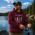 A man wearing a burgundy hoodie with a funny fishing-themed text graphic stands on a boat holding a fishing rod in front of a scenic lake and pine forest background.