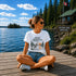 Woman sitting on a wooden dock by a lake with a cabin in the background, wearing a 'Still Waters' t-shirt.