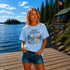 Woman standing on a dock at Falcon Lake Manitoba wearing a Still Waters heron graphic t-shirt featuring a heron, pine trees, calm lake water, and sunset artwork.