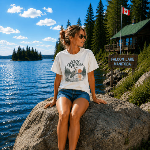Woman sitting on a rock at Falcon Lake Manitoba wearing a Still Waters heron graphic t-shirt featuring a heron, pine trees, calm lake water, and sunset artwork.