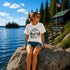 Woman sitting on a rock at Falcon Lake Manitoba wearing a Still Waters heron graphic t-shirt featuring a heron, pine trees, calm lake water, and sunset artwork.