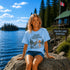 Woman sitting on a dock at Falcon Lake Manitoba wearing a Still Waters heron graphic t-shirt featuring a heron, pine trees, calm lake water, and sunset artwork.