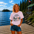 Woman standing on a dock at Falcon Lake Manitoba wearing a Still Waters heron graphic t-shirt featuring a heron, pine trees, calm lake water, and sunset artwork.
