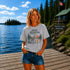 Woman standing on a dock at Falcon Lake Manitoba wearing a Still Waters heron graphic t-shirt featuring a heron, pine trees, calm lake water, and sunset artwork.