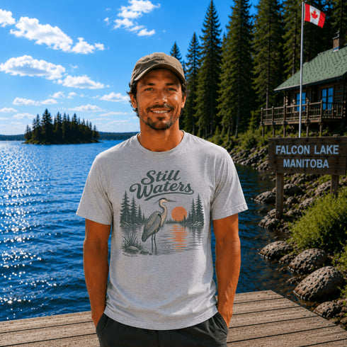 Man standing on a dock at Falcon Lake Manitoba wearing a Still Waters heron graphic t-shirt featuring a heron, pine trees, calm lake water, and sunset artwork.