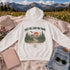 A white pullover hoodie laid flat on a pink blanket in a mountain meadow setting, with a large scenic graphic on the back reading “TAKE THE LONG WAY HOME” and “Every mile tells a story.” Surrounding props include a beige cap, a notebook, sunglasses, a bottle, and denim shorts.