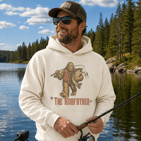 A man wearing a cream-colored hoodie with a graphic of a Bigfoot-style figure fishing and the text 'THE RODFATHER' stands by a lake with trees in the background.
