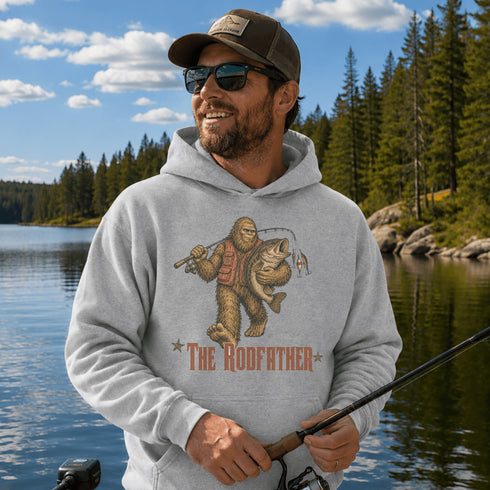 A man wearing a light gray hoodie with a 'The Rodfather' Bigfoot fishing graphic, standing on a boat by a lake with a fishing rod.