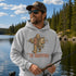 A man wearing a light gray hoodie with a 'The Rodfather' Bigfoot fishing graphic, standing on a boat by a lake with a fishing rod.