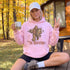A woman sitting outdoors on a wooden deck in front of a cabin, wearing a light pink hoodie with a large 'The Rodfather' graphic of a Bigfoot figure holding a fishing rod and reel. She is holding a white ceramic mug and making peace signs with both hands.