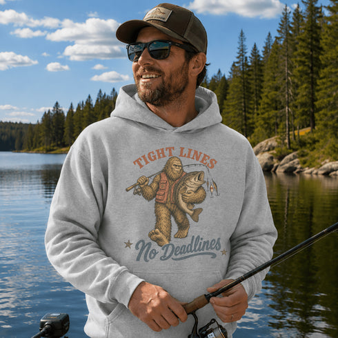 A man wearing a light gray hoodie with a 'Tight Lines No Deadlines' fishing graphic, standing on a lake with a forest background.