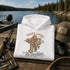 A folded white hoodie with a graphic print of a Bigfoot character fishing and the text 'TIGHT LINES No Deadlines' is displayed on a wooden dock by a lake. A fishing rod, tackle box, a brown trucker hat reading 'LIFE IS THE LAKE', and a black mug are arranged around it.