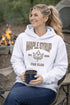 Woman wearing beige Maple Syrup Fan Club hoodie with maple leaf graphic, smiling outdoors in a natural setting with trees in the background