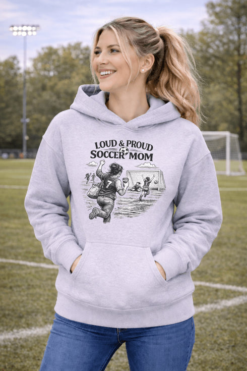 Woman wearing a grey Loud & Proud Soccer Mom hoodie with a vintage soccer scene design, standing on a soccer field with hands in pocket