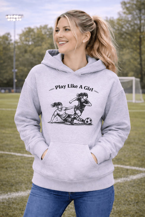 Woman wearing a grey Play Like A Girl soccer hoodie with girls soccer graphic, standing on a soccer field with hands in pocket