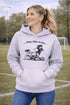 Woman wearing a grey Play Like A Girl soccer hoodie with girls soccer graphic, standing on a soccer field with hands in pocket