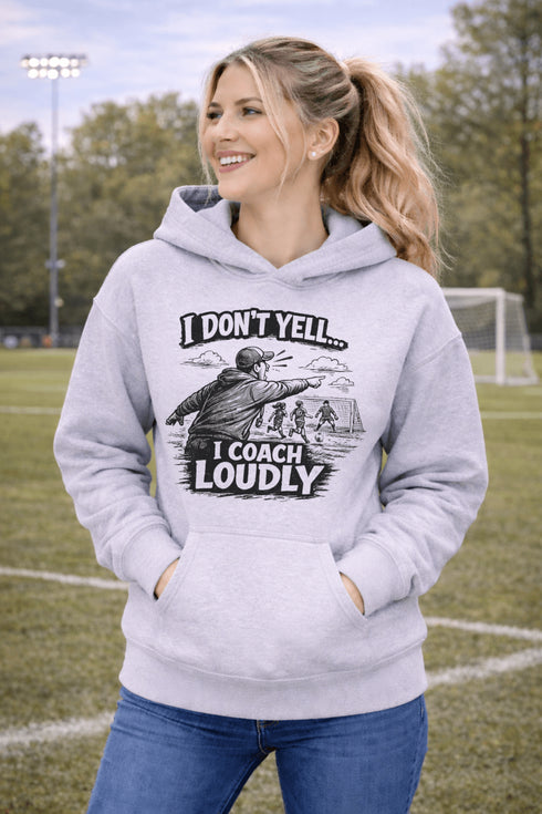 Woman wearing a grey hoodie with “I Don’t Yell I Coach Loudly” soccer coach design featuring coach pointing on sideline with players in background