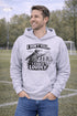 man wearing a grey hoodie with “I Don’t Yell I Coach Loudly” soccer coach design featuring coach pointing on sideline with players in background
