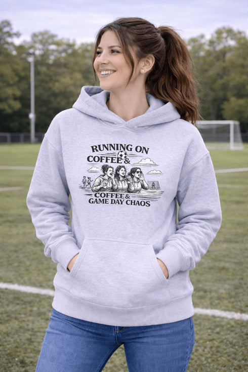 Soccer mom hoodie with “Running on Coffee and Game Day Chaos” design featuring moms cheering on the sidelines with kids playing soccer in the background