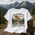 White t-shirt with 'Somewhere Between Lost & Free' design on a wooden surface with a mountainous background