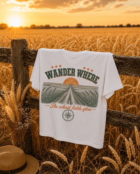 White t-shirt with 'Wander Where' design hanging on a wooden fence in a wheat field at sunset.