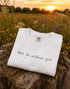 Folded white t-shirt with 'Where the wildflowers grow' text on a wooden stump with a sunset in the background.