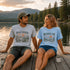 Two people sitting on a dock by a lake, wearing t-shirts with nature-themed designs.