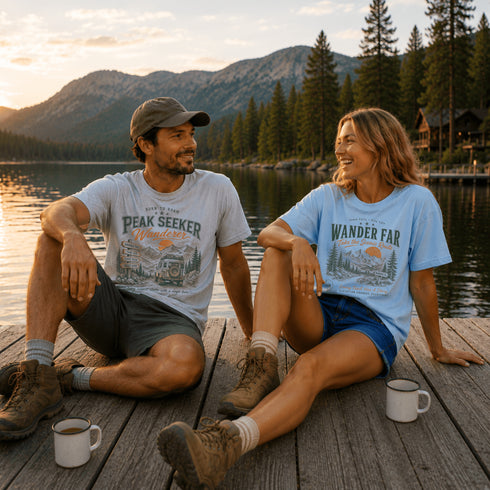Two people sitting on a dock by a lake with mountains in the background, wearing t-shirts with nature-themed designs.