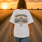 Woman wearing a white Comfort Colors t-shirt featuring a vintage prairie road and wheat field sunset graphic on the back while standing on a gravel road surrounded by golden wheat fields at sunset.