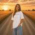 Woman wearing a white Comfort Colors t-shirt featuring a vintage prairie road and wheat field sunset graphic on the back while standing on a gravel road surrounded by golden wheat fields at sunset.