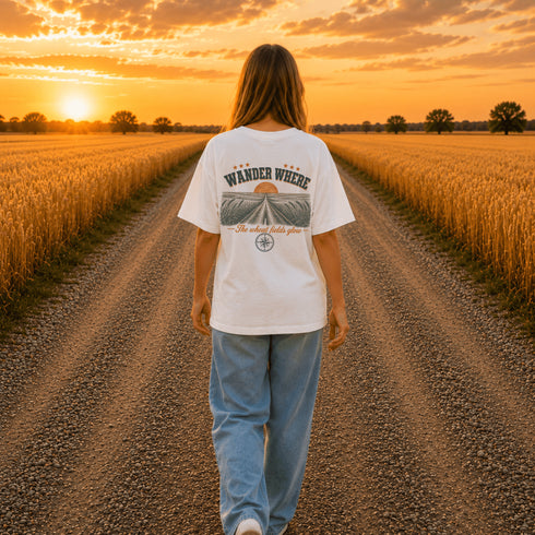 Woman wearing a white Comfort Colors t-shirt featuring a vintage prairie road and wheat field sunset graphic on the back while standing on a gravel road surrounded by golden wheat fields at sunset.