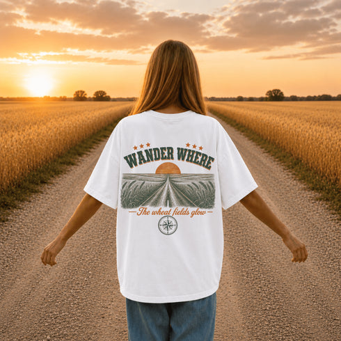 Woman wearing a white Comfort Colors t-shirt featuring a vintage prairie road and wheat field sunset graphic on the back while standing on a gravel road surrounded by golden wheat fields at sunset.