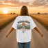 Woman wearing a white Comfort Colors t-shirt featuring a vintage prairie road and wheat field sunset graphic on the back while standing on a gravel road surrounded by golden wheat fields at sunset.
