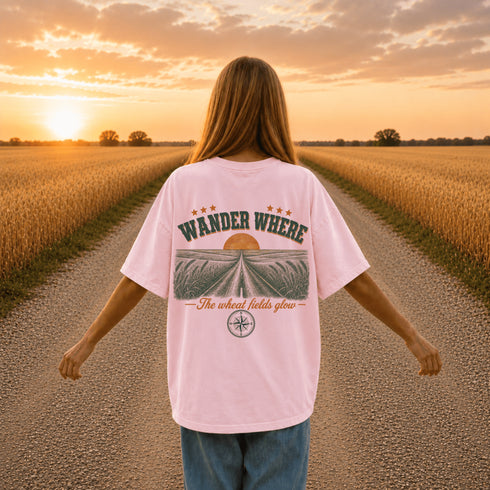 Woman wearing a white Comfort Colors t-shirt featuring a vintage prairie road and wheat field sunset graphic on the back while standing on a gravel road surrounded by golden wheat fields at sunset.