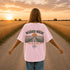 Woman wearing a white Comfort Colors t-shirt featuring a vintage prairie road and wheat field sunset graphic on the back while standing on a gravel road surrounded by golden wheat fields at sunset.