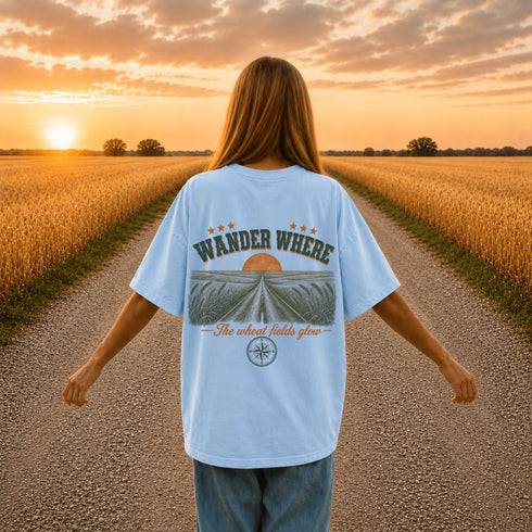 Woman wearing a white Comfort Colors t-shirt featuring a vintage prairie road and wheat field sunset graphic on the back while standing on a gravel road surrounded by golden wheat fields at sunset.