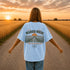 Woman wearing a white Comfort Colors t-shirt featuring a vintage prairie road and wheat field sunset graphic on the back while standing on a gravel road surrounded by golden wheat fields at sunset.