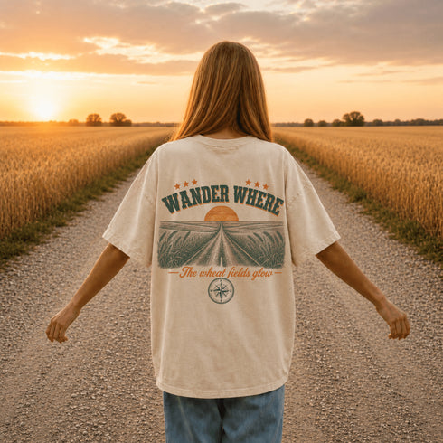 Woman wearing a white Comfort Colors t-shirt featuring a vintage prairie road and wheat field sunset graphic on the back while standing on a gravel road surrounded by golden wheat fields at sunset.