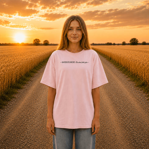 Woman wearing a white Comfort Colors t-shirt featuring a vintage prairie road and wheat field sunset graphic on the back while standing on a gravel road surrounded by golden wheat fields at sunset.