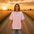 Woman wearing a white Comfort Colors t-shirt featuring a vintage prairie road and wheat field sunset graphic on the back while standing on a gravel road surrounded by golden wheat fields at sunset.
