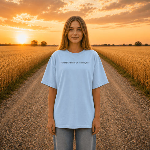 Woman wearing a white Comfort Colors t-shirt featuring a vintage prairie road and wheat field sunset graphic on the back while standing on a gravel road surrounded by golden wheat fields at sunset.